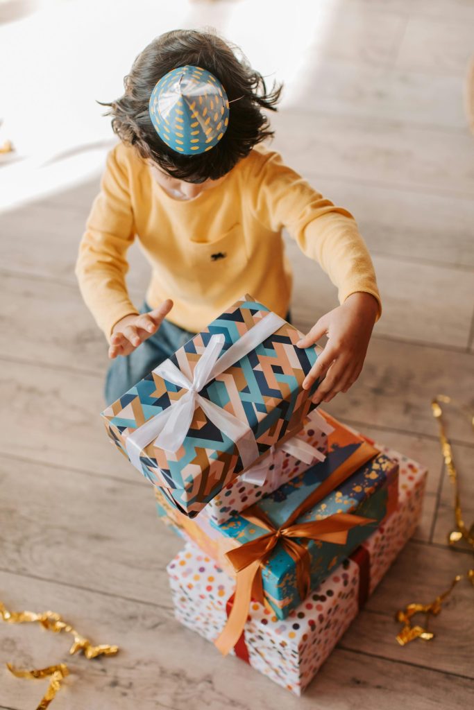 A young boy celebrates with a stack of colorful presents on a wooden floor.